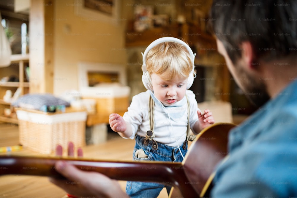 Jeune père méconnaissable à la maison jouant de la guitare, son petit fils avec des écouteurs, écoutant de la musique et dansant.