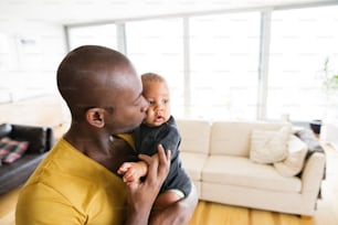 自宅で若いアフリカ系アメリカ人の父親がかわいい息子を腕に抱き、頬にキスをしている。