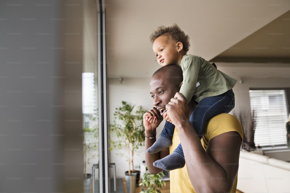 Junger afroamerikanischer Vater zu Hause am Fenster und trägt seine kleine Tochter auf den Schultern.