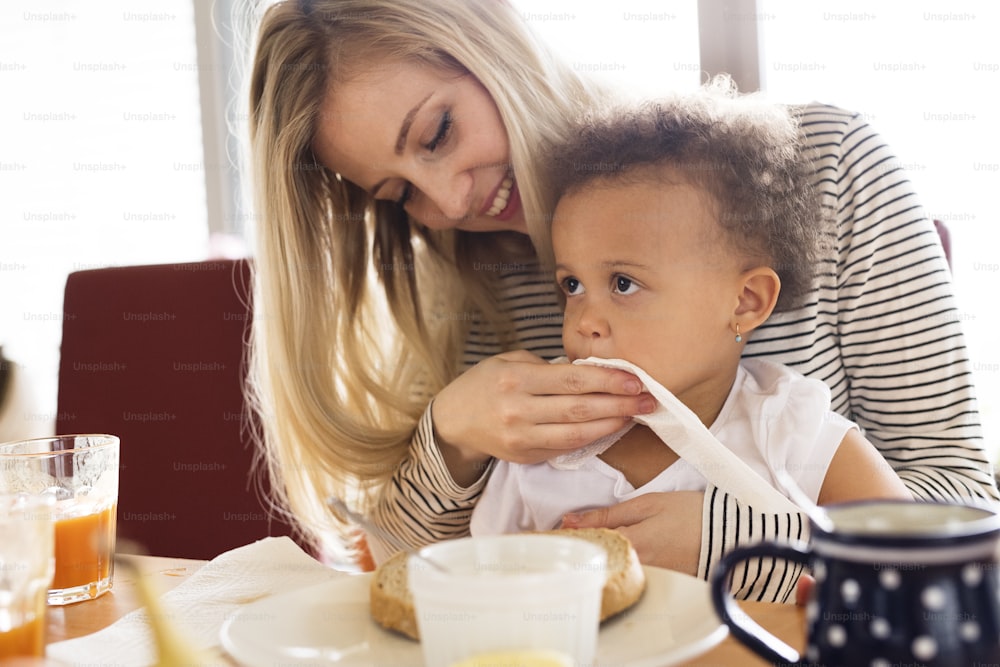 Beautiful young mother at home with his little mixed-race daughter sitting on her lap, having breakfast together.