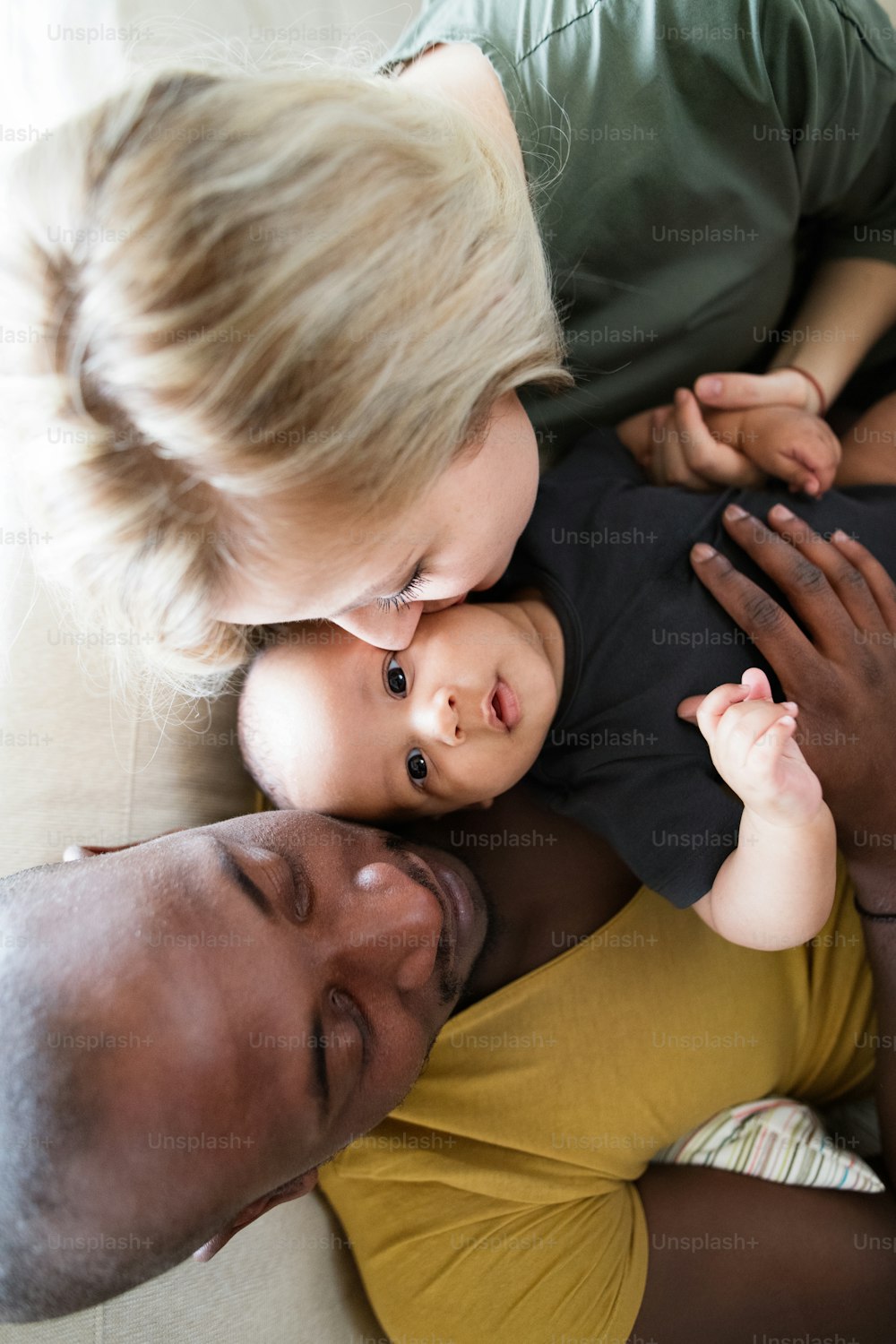 Beautiful young interracial family at home holding their cute little baby son, mother kissing him on cheek.