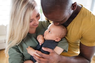 Hermosa familia interracial joven en casa sosteniendo a su lindo bebé, padre besándolo en la frente.