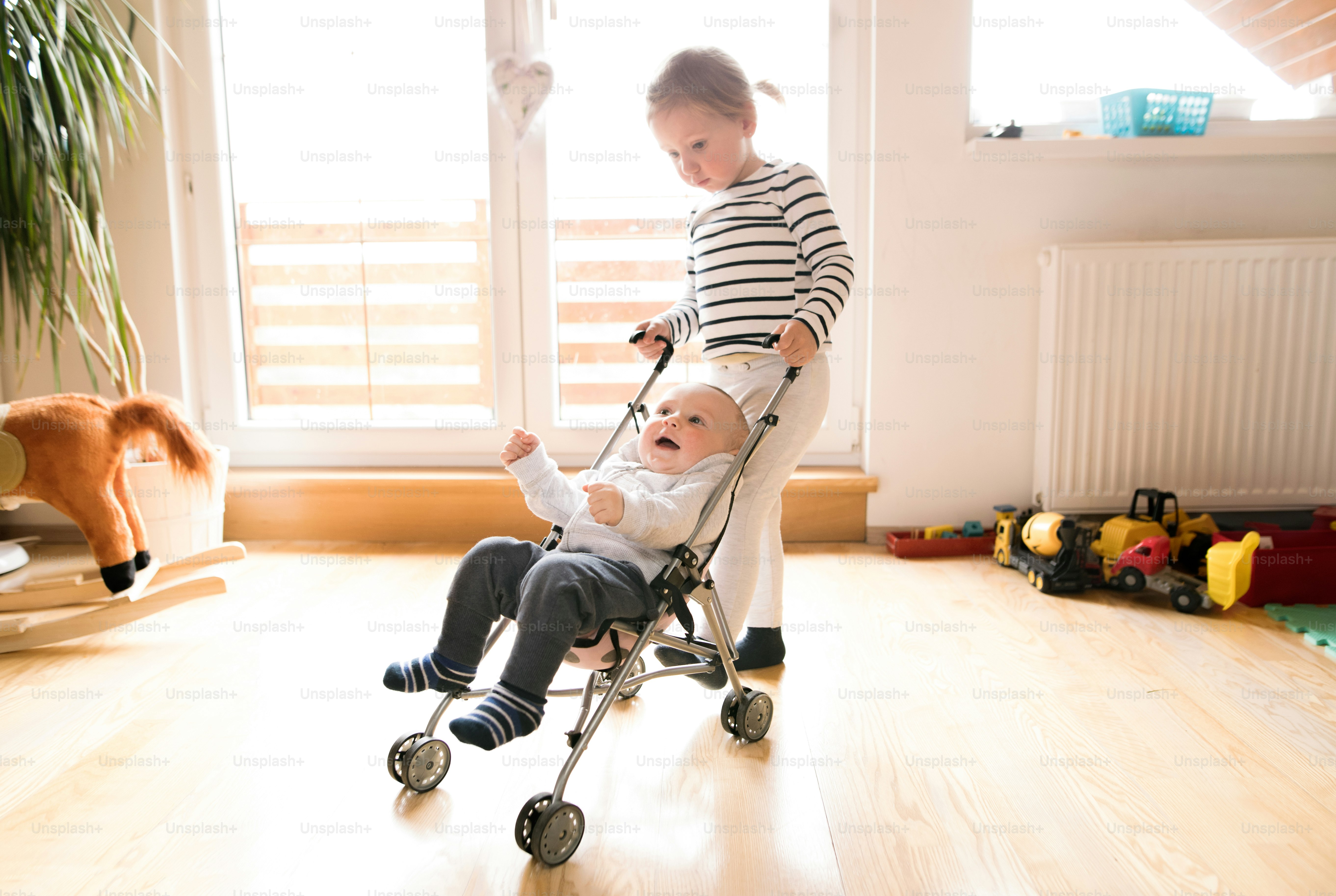 Cute little girl at home with her baby brother in toy stroller, playing with him.