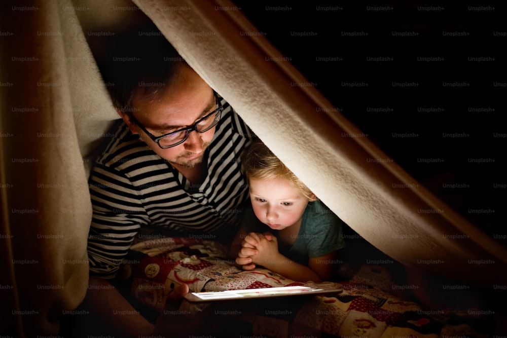 Padre joven en casa con su linda hijita bajo manta por la noche viendo algo en tableta.