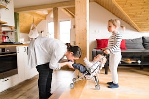 Cute little girl at home with her baby brother in toy stroller, playing with him, his mother kissing his feet.