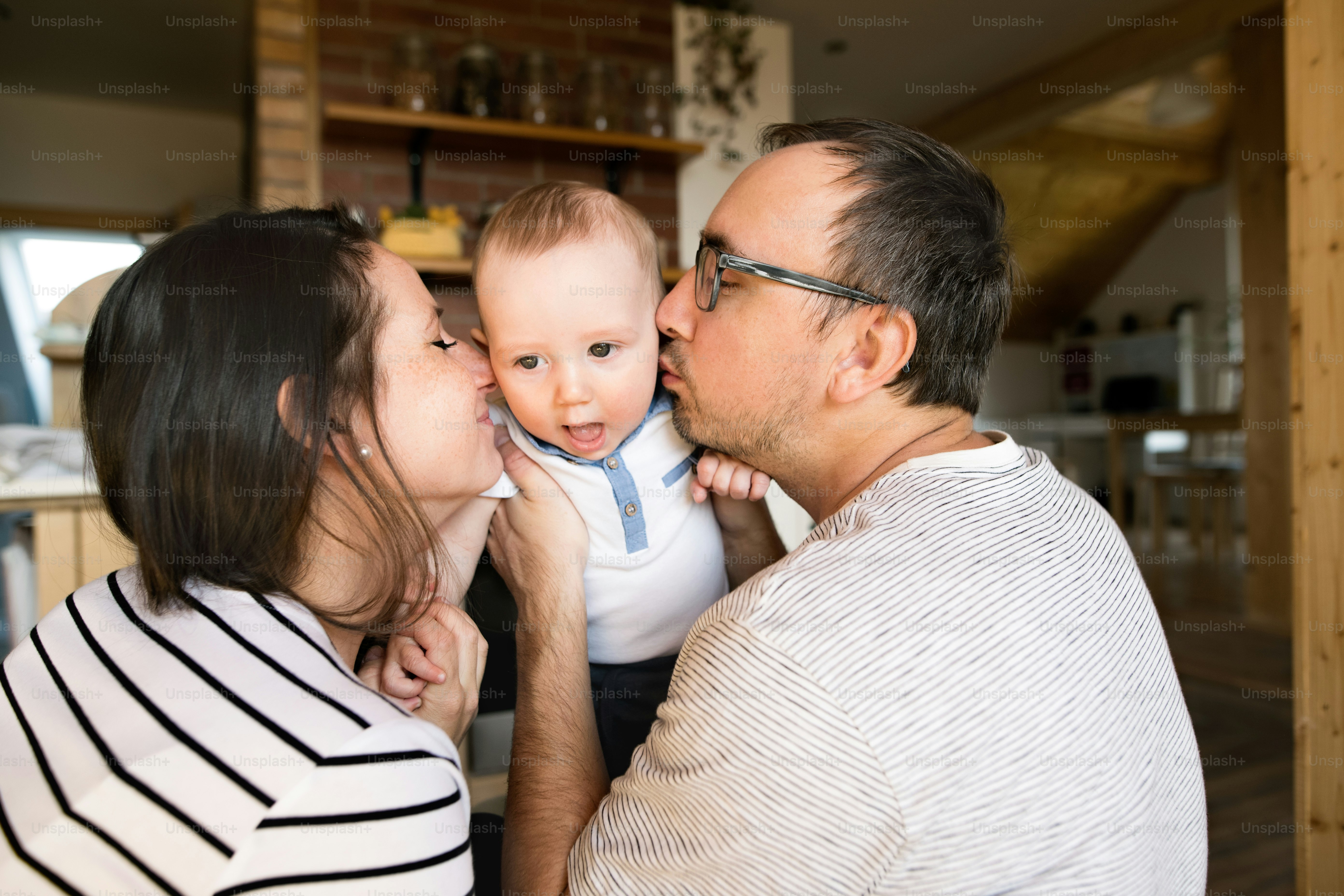 Beautiful young parents at home holding their cute baby son in the arms, kissing him on cheeks.