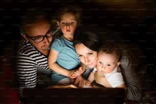 Giovani genitori a casa di notte con i loro bambini piccoli che guardano qualcosa sul computer portatile.