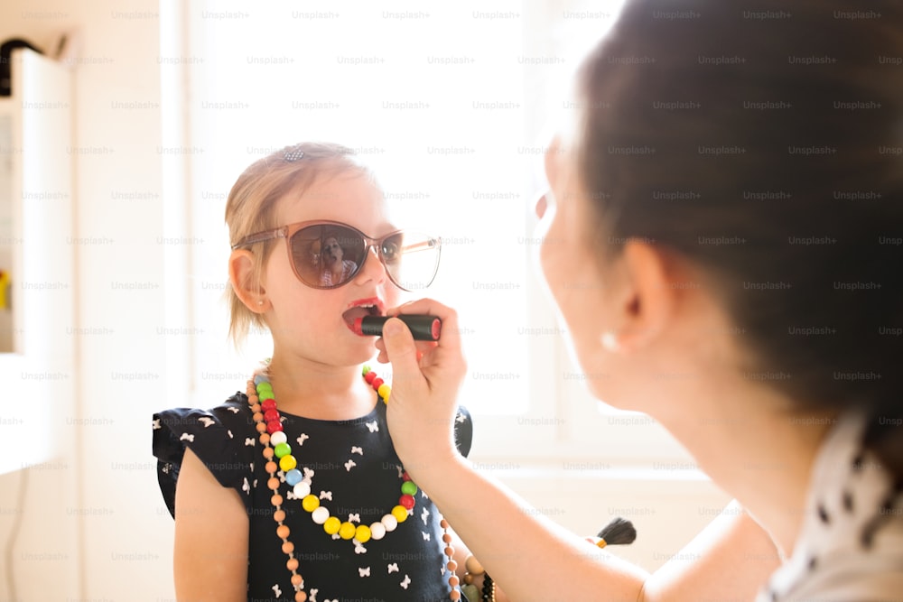 Unrecognizable young mother painting lips of her cute little daughter with red lipstick.