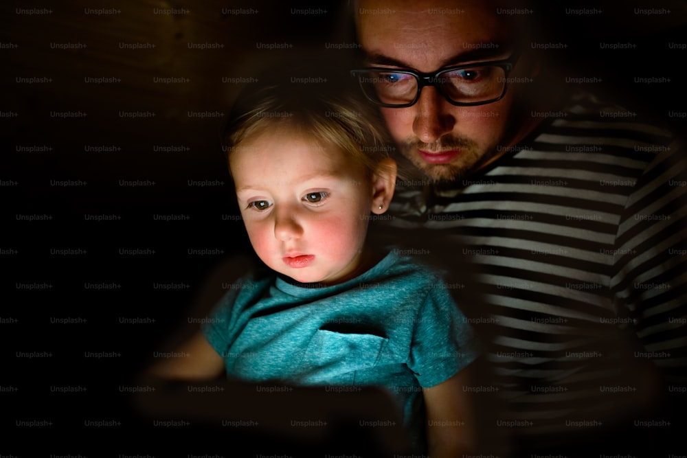 Giovane padre a casa di notte con la sua graziosa figlioletta sulle ginocchia a giocare o guardare qualcosa su tablet.