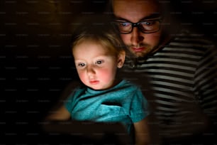 Young father at home at night with his cute little daughter on his lap playing or watching something on tablet.