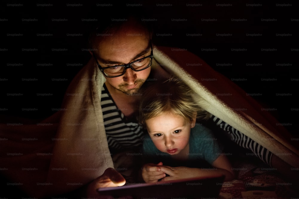 젊은 아버지는 밤에 담요 아래 귀여운 딸과 함께 태블릿으로 무언가를 보고 있습니다.