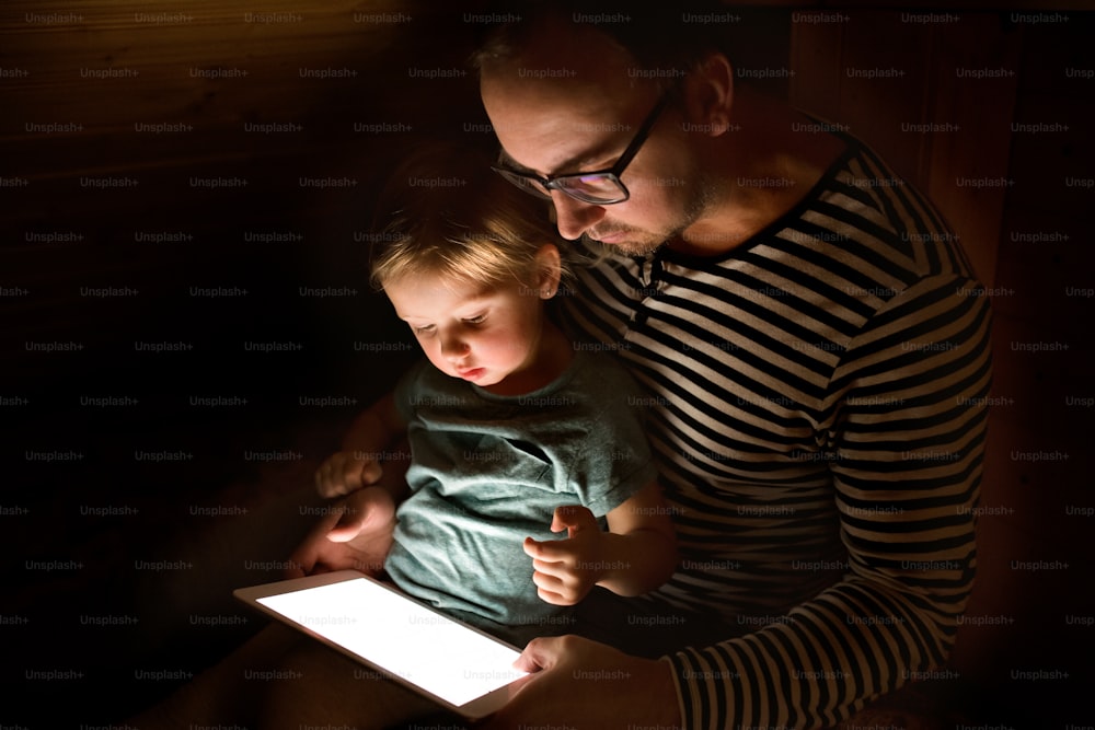 Young father at home at night with his cute little daughter on his lap playing or watching something on tablet.
