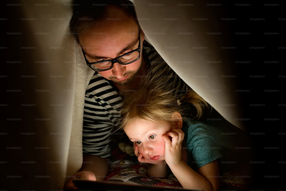Jovem pai em casa com sua filhinha fofa debaixo do cobertor à noite assistindo algo no tablet.