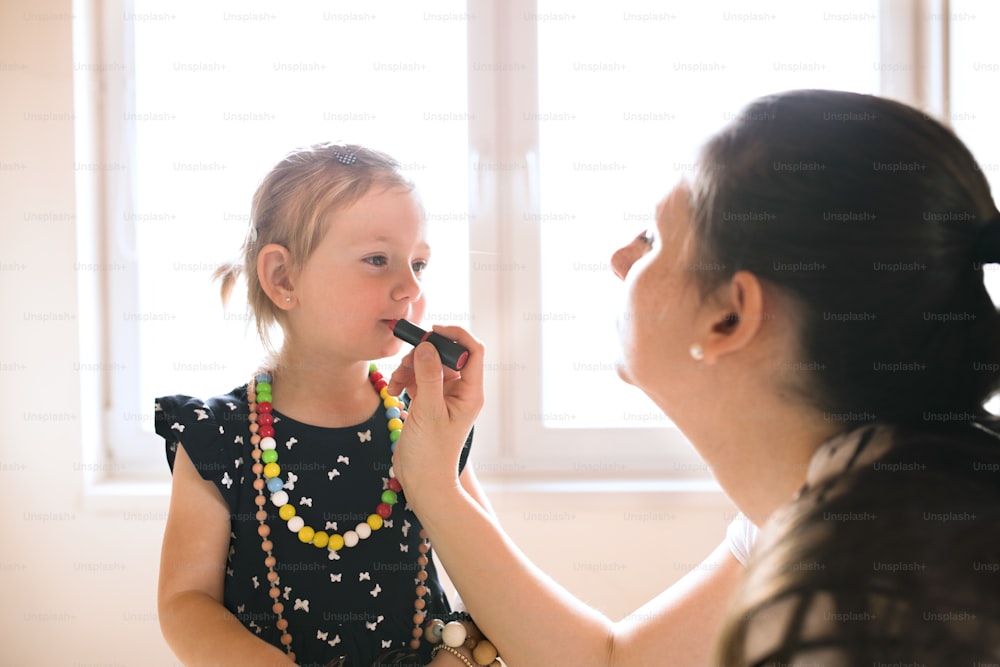 Hermosa joven madre pintando los labios de su linda hijita con lápiz labial.