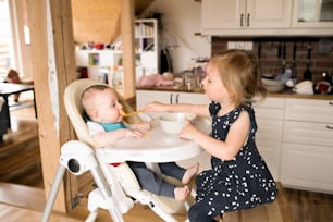 Dos niños pequeños en casa, linda niña alimentando a su hermanito sentado en silla alta.