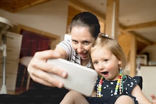 Beautiful young mother at home and her cute little daughter taking selfie with smart phone.