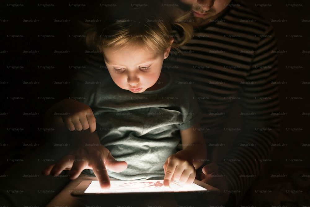 Pai irreconhecível em casa à noite com sua filhinha fofa no colo brincando ou assistindo algo no tablet.