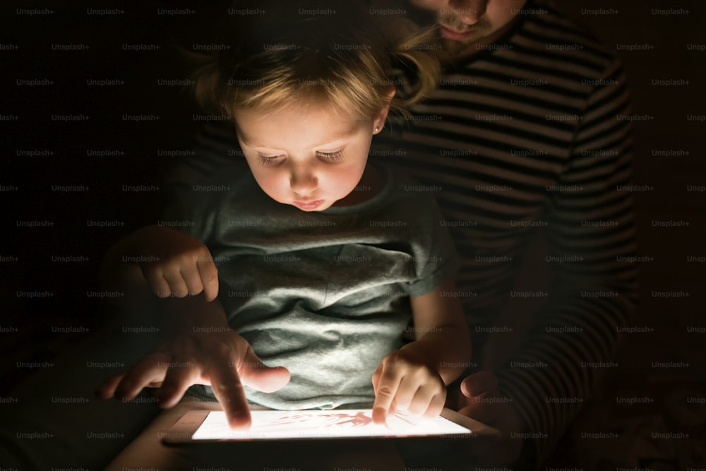 밤에 집에서 알아볼 수 없는 아버지는 귀여운 딸을 무릎에 앉히고 태블릿에서 무언가를 보거나 놀고 있습니다.