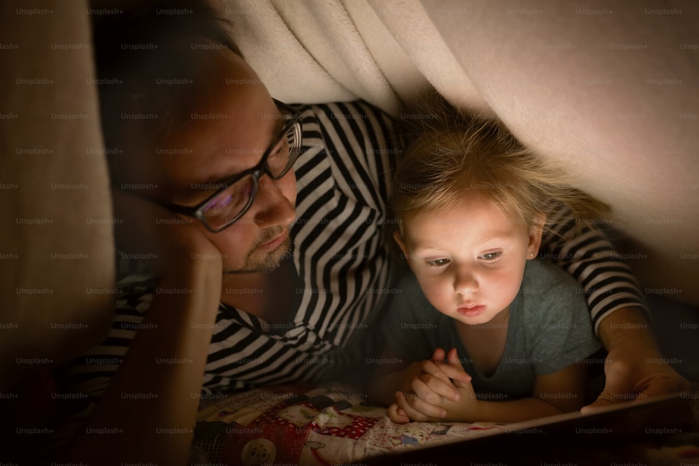 Padre joven en casa por la noche con su linda hijita jugando o viendo algo en la tableta.