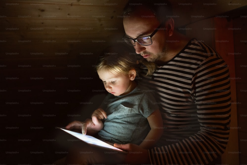 Giovane padre a casa di notte con la sua graziosa figlioletta sulle ginocchia a giocare o guardare qualcosa su tablet.