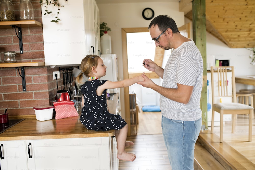 Padre joven en casa en la cocina con su linda hijita pintándose las uñas.