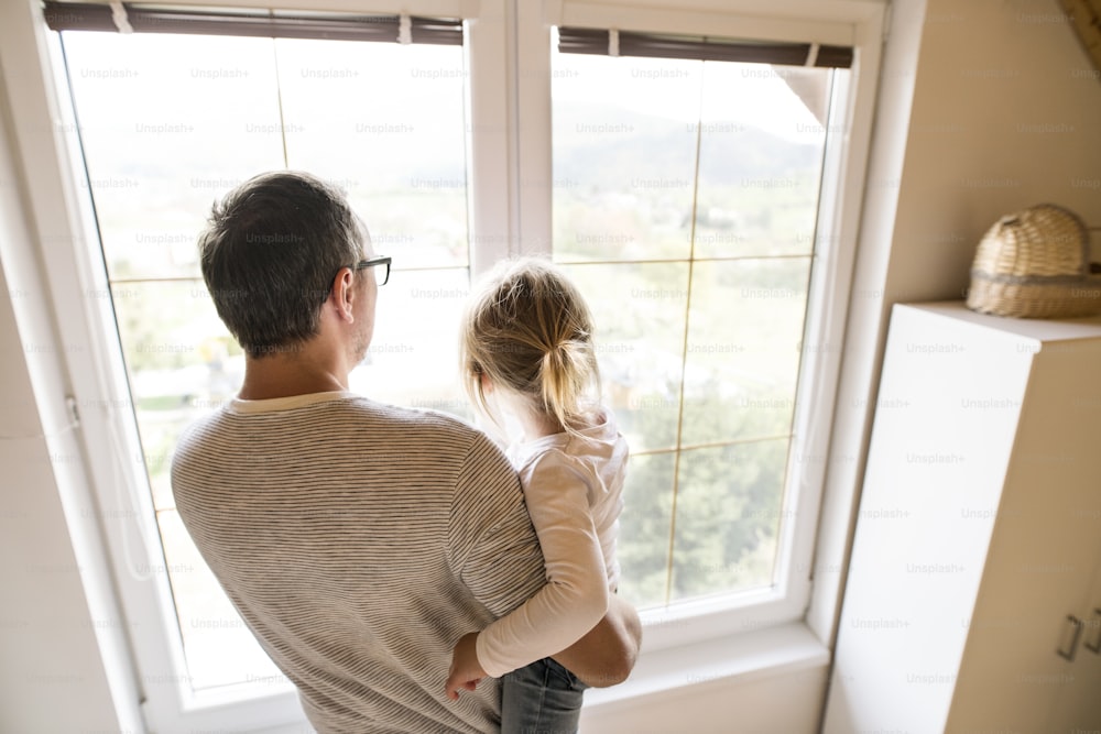 귀여운 딸을 품에 안고 창가에 서 있는 집에서 알아볼 수 없는 젊은 아버지.