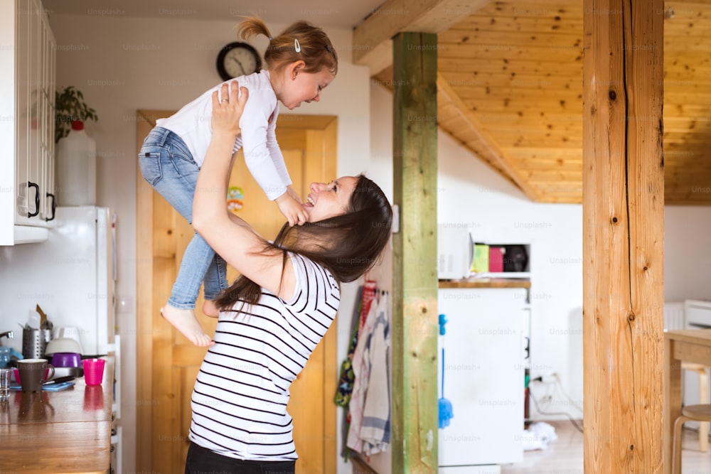 Linda jovem mãe em casa na cozinha levantando sua filhinha fofa no ar.