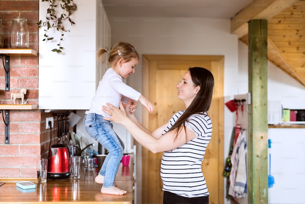 Linda jovem mãe em casa e sua filhinha fofa pulando da bancada da cozinha para seus braços.