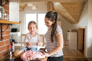 Schöne junge Mutter zu Hause und ihre süße kleine Tochter sitzt auf Küchenarbeitsplatte und spielt mit Puppe.