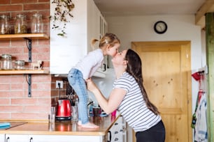 Beautiful young mother at home and her cute little daughter standing on kitchen countertop kissing her