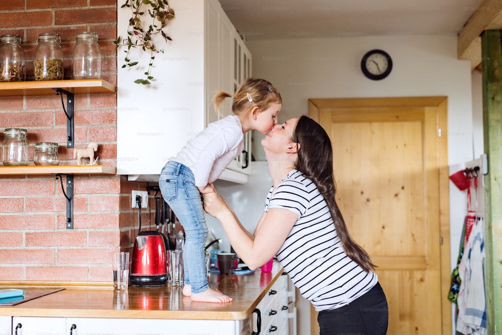 Bela jovem mãe em casa e sua filhinha fofa em pé na bancada da cozinha beijando-a