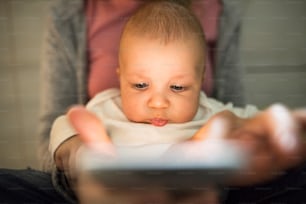 Unrecognizable young mother at home with her little baby son in the arms, sitting on the kitchen floor, holding smart phone