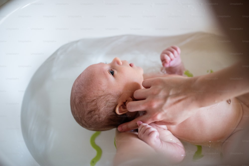 Mãe irreconhecível segurando seu filho bebê, banhando-o em um pequeno banho de plástico branco. De perto.