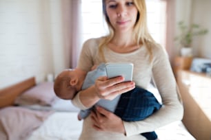 Beautiful young mother holding her sleeping baby son in her arms white sending text message from her smart phone