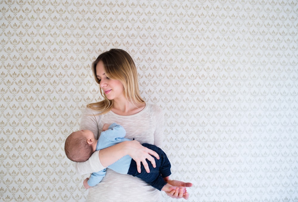 Beautiful young mother holding her sleeping baby son in her arms standing in front of wall with wallpaper on it