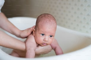 Unrecognizable mother holding her baby son, bathing him in small white plastic bath. Close up.