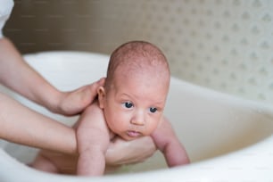 Madre irreconocible sosteniendo a su bebé, bañándolo en un pequeño baño de plástico blanco. Cerrar.
