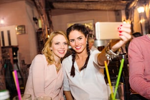Jeunes belles femmes avec des cocktails dans un bar ou un club en prenant des selfies, en s’amusant