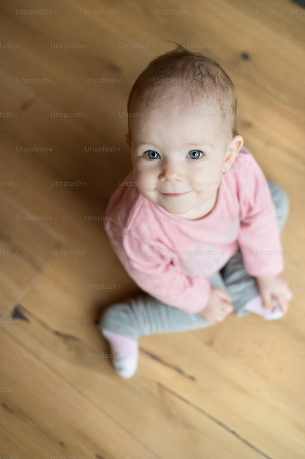 Cute little girl in pink sweatshirt at home sitting on wooden floor, looking up, smiling