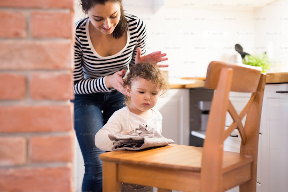 Bela jovem mãe com sua filha pequena bonito na cozinha, menina limpando cadeira de madeira com pano de prato