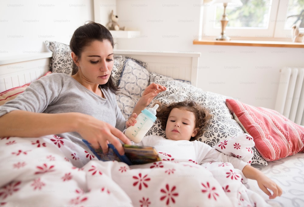 Linda jovem mãe deitada na cama, colocando sua filhinha fofa para dormir, lendo seu livro.