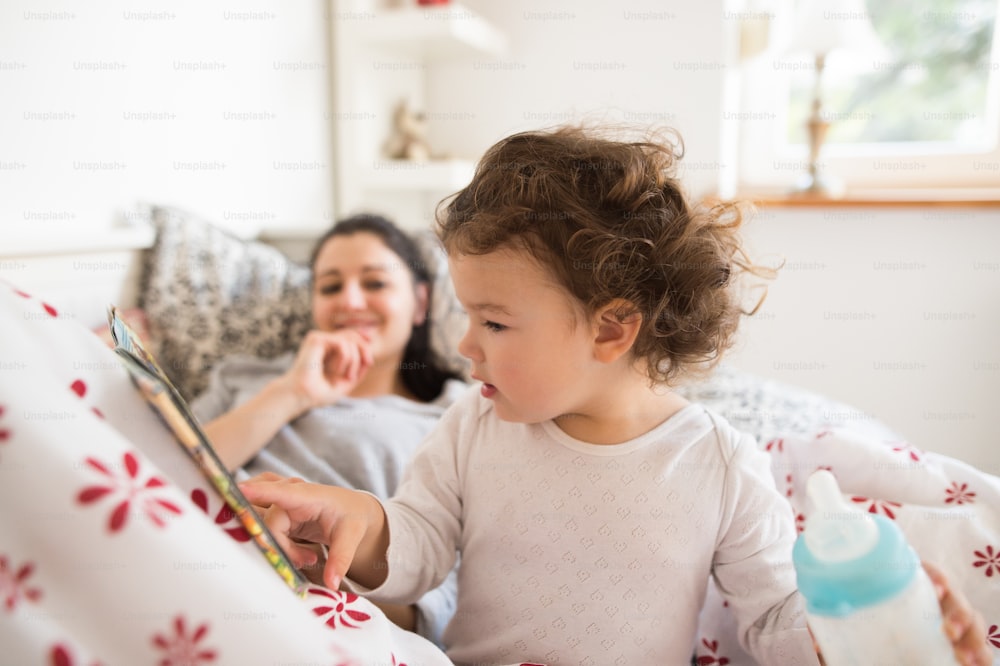 Beautiful young mother lying in bed, putting her cute little daughter to sleep, girl reading her book.