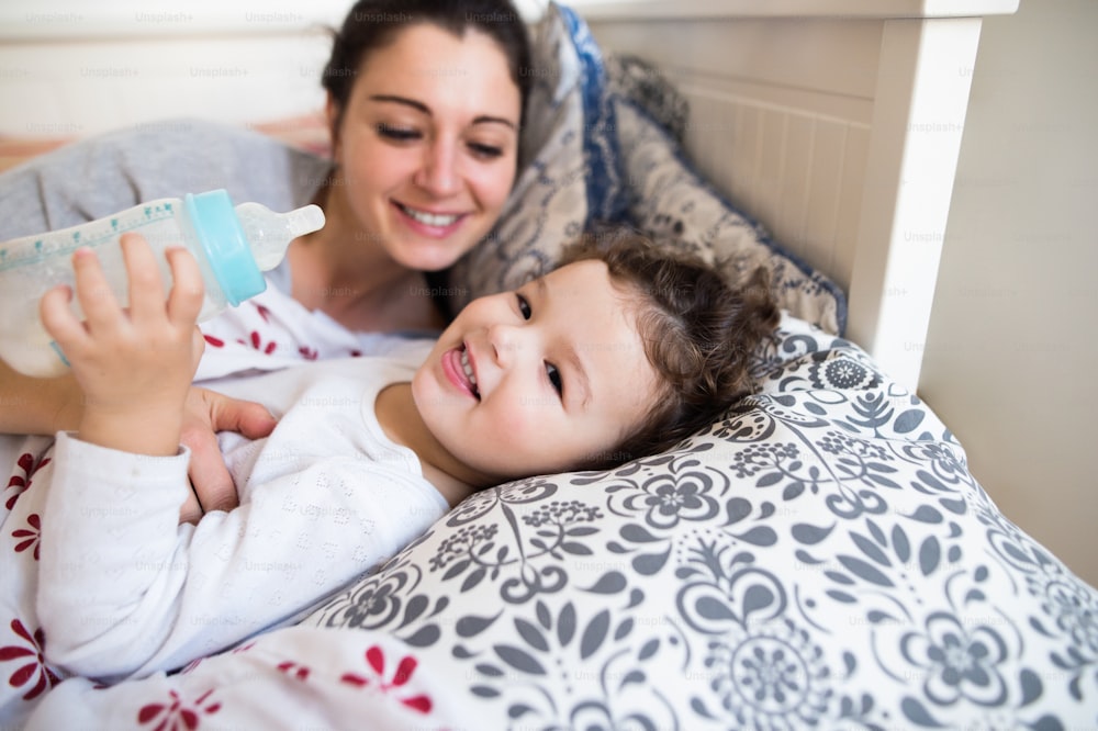 아름다운 젊은 어머니가 침대에 누워 귀여운 딸을 재우고, 우유병을 들고 있는 소녀.