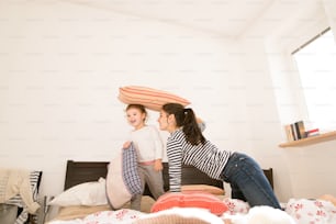 Hermosa madre joven divirtiéndose con su linda hija en la cama de su habitación, jugando con almohadas