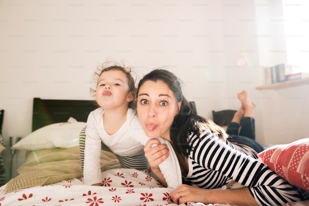 Beautiful young mother having fun with her cute little daughter on bed in her bedroom, sticking tongues out