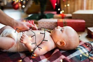 Padre irreconocible con bebé en Navidad. Bebé acostado sobre la manta en el suelo., enredado en la cadena de luces.