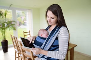 Schöne junge Mutter mit ihrem neugeborenen Sohn im Tragetuch zu Hause, hält Tablet, schreibt etwas