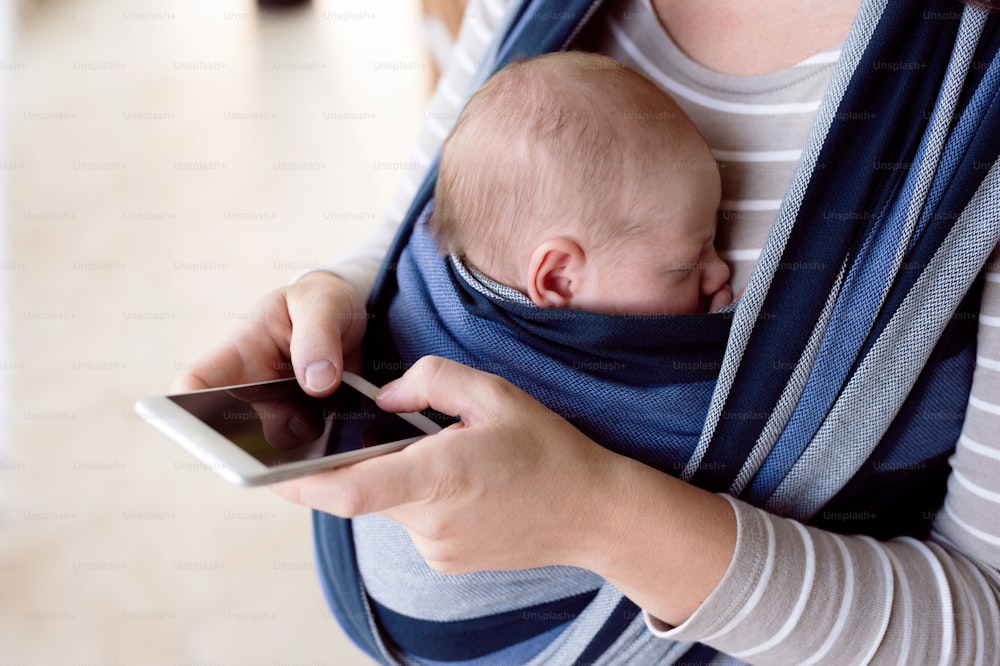 Madre joven irreconocible con su hijo recién nacido en cabestrillo en casa, sosteniendo un teléfono inteligente, escribiendo algo