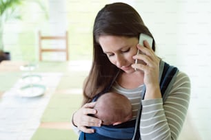 Beautiful young mother with her newborn baby son in sling at home, holding smart phone, making phone call