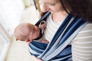 Close up, beautiful young mother carrying her newborn baby son in sling at home
